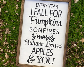 May include: A rectangular wooden framed sign with the text "EVERY YEAR I FALL FOR Pumpkins BONFIRES & s'mores Autumn Leaves APPLES & YOU" in black script on a white background. The sign is resting on a bed of green foliage.