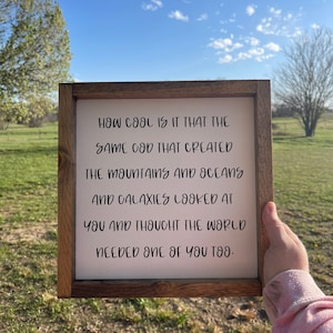 May include: A wooden framed sign with the text "HOW COOL IS IT THAT THE SAME GOD THAT CREATED THE MOUNTAINS AND OCEANS AND GALAXIES LOOKED AT YOU AND THOUGHT THE WORLD NEEDED ONE OF YOU TOO." The sign is held up against a blue sky.