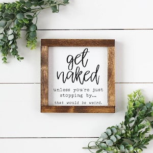May include: A square wooden sign with a white background and a dark brown frame. The sign reads "get naked" in a handwritten font, followed by "unless you're just stopping by... that would be weird." Greenery surrounds the sign.
