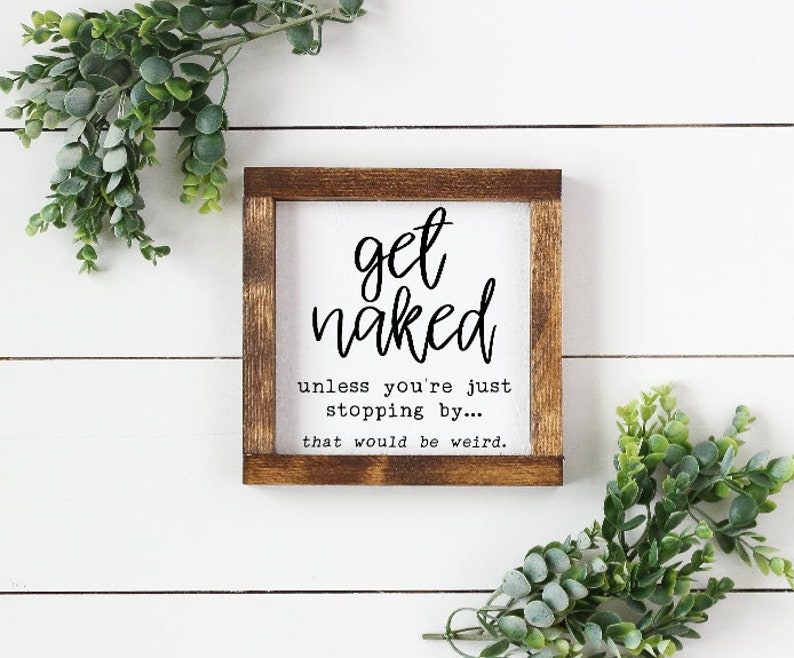 May include: A square wooden sign with a white background and a dark brown frame. The sign reads "get naked" in a handwritten font, followed by "unless you're just stopping by... that would be weird." Greenery surrounds the sign.