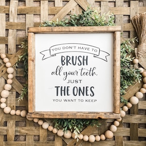 May include: A framed sign with the text "YOU DON'T HAVE TO BRUSH all your teeth, JUST THE ONES YOU WANT TO KEEP." The sign is surrounded by greenery and wooden beads, set against a woven background.
