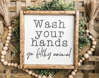May include: A wooden framed sign with the text "Wash your hands ya filthy animal" in black lettering. The sign is surrounded by greenery and a wooden bead garland, set against a woven basket background.