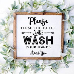 May include: A square wooden sign with the text "Please FLUSH THE TOILET AND WASH YOUR HANDS Thank You" in black lettering. The sign is surrounded by a garland of green and gray leaves.