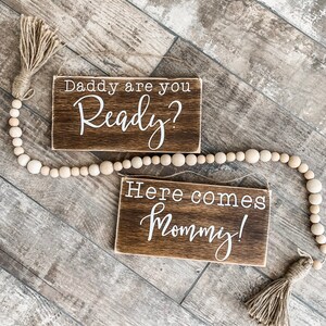 May include: Two small, rectangular wooden signs with white lettering. The top sign reads "Daddy are you Ready?" and the bottom sign says "Here comes Mommy!". A beaded garland and tassels are also present.