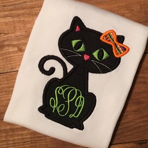 May include: A white t-shirt with an embroidered black cat wearing a bright orange and green bow. The cat has green eyes and a pink nose. The cat's tail is curled. The cat's body has a green monogram.