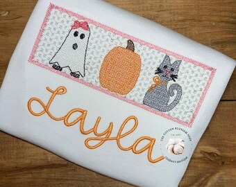 May include: A white onesie with a pink, orange, and gray embroidered design of a ghost, pumpkin, and cat. The name "Layla" is embroidered in orange below the design. The text "The Cotton Blossom Shop Est. 2022 Children's Boutique" is embroidered in white on a small white cotton ball in the bottom right corner.