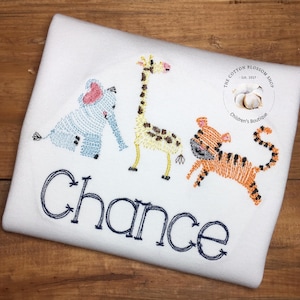 Puede incluir: Un pelele blanco para bebé con animales bordados, incluyendo un elefante, una jirafa y un tigre. El pelele tiene el nombre "Chance" bordado en azul debajo de los animales. El pelele tiene un pequeño logotipo en la esquina superior derecha que dice "The Cotton Blossom Shop - Est. 2017 - Children's Boutique."