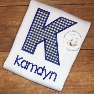 May include: White onesie with a blue and white gingham letter K and the name "Kamdyn" embroidered in blue. The onesie has a small logo that says "The Cotton Blossom Shop - Est. 2017 - Children's Boutique".