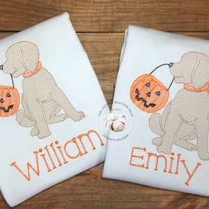 May include: Two white t-shirts with embroidered images of a dog holding a jack-o'-lantern. The shirts are personalized with the names "William" and "Emily".