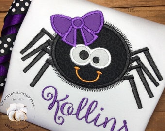 May include: A white onesie with a black and purple embroidered spider wearing a purple bow. The onesie is personalized with the name "Rollins" in purple cursive.