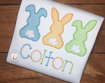 May include: A white onesie with three embroidered bunnies in blue, yellow, and green. The bunnies have white feet and tails. The name "Colton" is embroidered below the bunnies in a colorful font.
