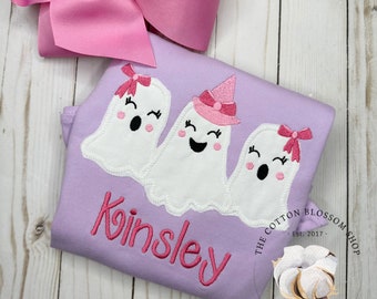 May include: A lavender-colored shirt with three embroidered white ghosts wearing pink bows and hats. The shirt has the name "Kinsley" embroidered in pink on the front. The shirt also has a small embroidered logo that says "The Cotton Blossom Shop Est. 2017" and "Children's Boutique".