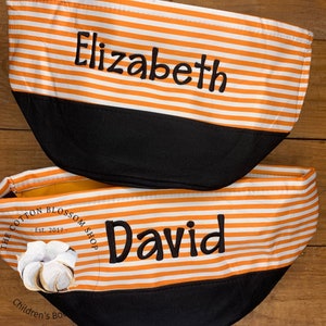 May include: Two orange and white striped fabric storage bins with black bottoms. The top bin has the name "Elizabeth" embroidered on it. The bottom bin has the name "David" embroidered on it. The bin has a black and white logo with the text "The Cotton Blossom Shop - Est. 2017 - Children's Boutique".