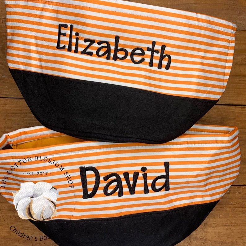 May include: Two orange and white striped fabric storage bins with black bottoms. The top bin has the name "Elizabeth" embroidered on it. The bottom bin has the name "David" embroidered on it. The bin has a black and white logo with the text "The Cotton Blossom Shop - Est. 2017 - Children's Boutique".