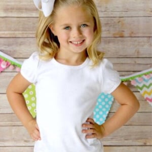 May include: A young girl wearing a white short-sleeved t-shirt with a white bow in her hair. She is standing in front of a wooden background with colourful decorations.