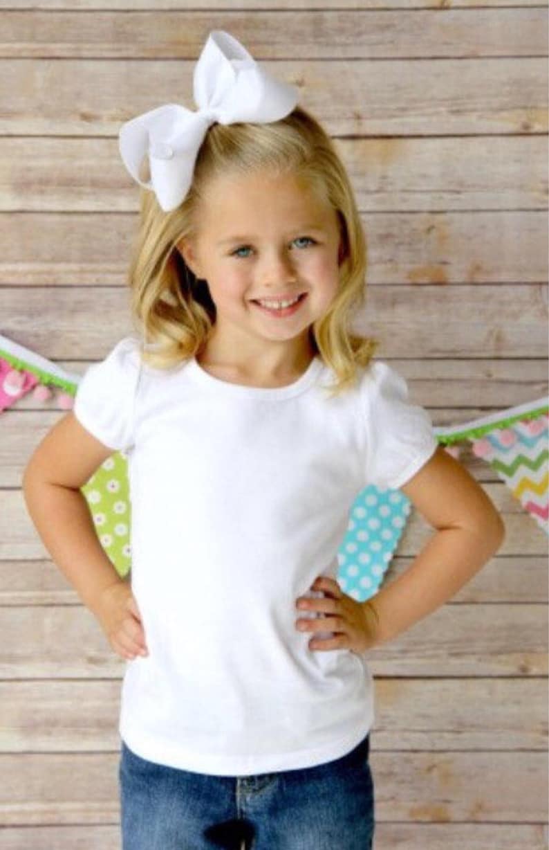 May include: A young girl wearing a white short-sleeved t-shirt with a white bow in her hair. She is standing in front of a wooden background with colourful decorations.