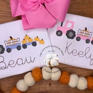 May include: Two white shirts with embroidered pumpkin patch designs. The shirts feature a blue and a pink tractor pulling a wagon full of pumpkins. The shirts are personalized with the names "Beau" and "Keely".