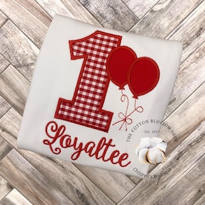 May include: Red and white gingham number one with two red balloons and the name Loyalttee embroidered on a white t-shirt. The Cotton Blossom Shop Children's Boutique logo is in the bottom right corner.