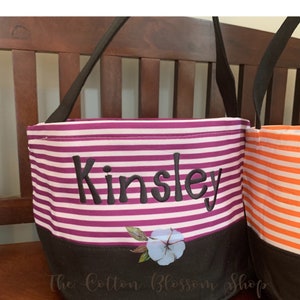 May include: Purple and white striped Halloween trick-or-treat bag with black handles and a black bottom. The bag has the name "Kinsley" embroidered on the front and a cotton blossom design on the bottom.