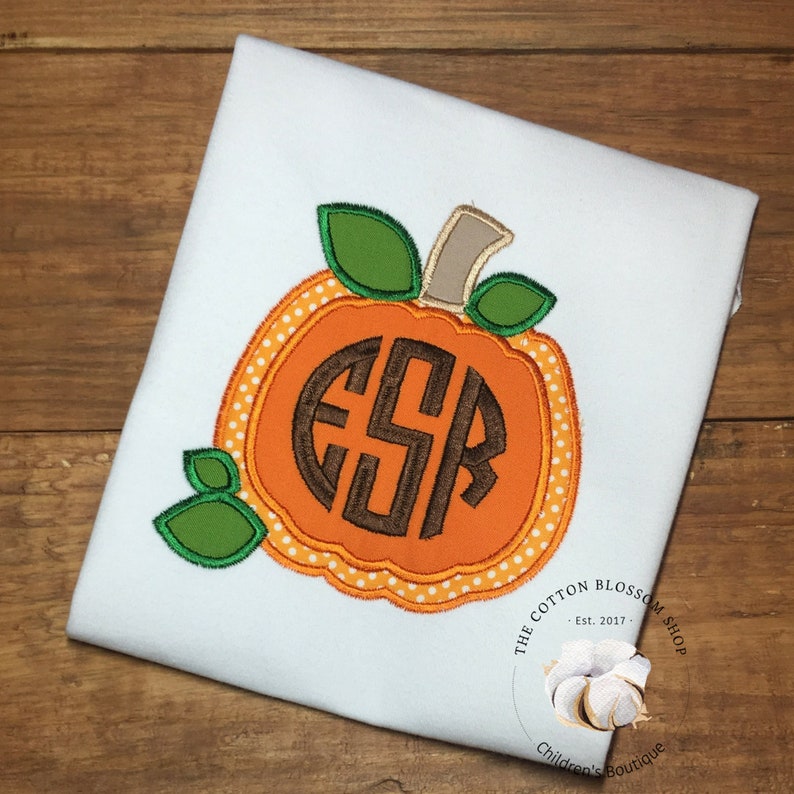 May include: A white t-shirt with an orange pumpkin applique. The pumpkin has a brown monogram "ESR" in the center and green leaves on the top. The text "The Cotton Blossom Shop - Est. 2017 - Children's Boutique" is printed on the bottom right corner of the shirt.