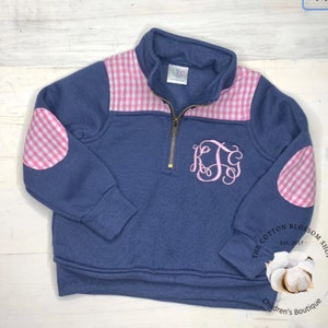 Puede incluir: Un jersey azul marino con cuello alzado con cremallera, con detalles de cuadros rosas y blancos en los hombros y coderas. La parte delantera muestra un monograma rosa. El logotipo de The Cotton Blossom Shop está en la esquina inferior derecha.