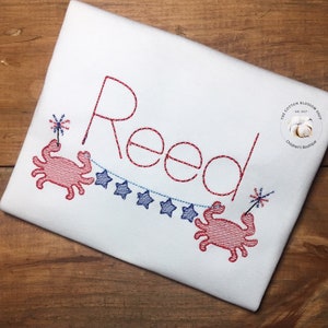 May include: White t-shirt with red embroidered name "Reed". Features two red crabs holding fireworks and a blue star banner. The Cotton Blossom Shop logo is in the upper right corner.
