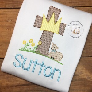 May include: A white embroidered t-shirt featuring a brown cross, a yellow crown, a white lamb, and green grass. The name "Sutton" is embroidered in blue below the cross. The t-shirt has a small white circle with the text "The Cotton Blossom Shop Est. 2017 Children's Boutique" in black.