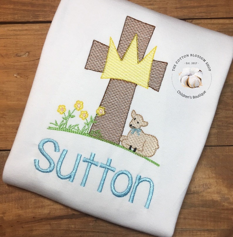 May include: A white embroidered t-shirt featuring a brown cross, a yellow crown, a white lamb, and green grass. The name "Sutton" is embroidered in blue below the cross. The t-shirt has a small white circle with the text "The Cotton Blossom Shop Est. 2017 Children's Boutique" in black.