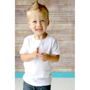 May include: A young boy with blonde hair styled into a mohawk wears a white t-shirt. He is smiling and looking at the camera.