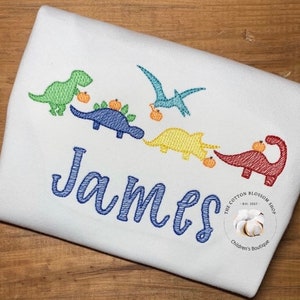 May include: A white onesie with a blue embroidered name "James" and five colorful dinosaurs carrying pumpkins. The dinosaurs are green, blue, yellow, and red. The onesie has a small logo in the bottom right corner that says "The Cotton Blossom Shop - Est. 2017 - Children's Boutique."