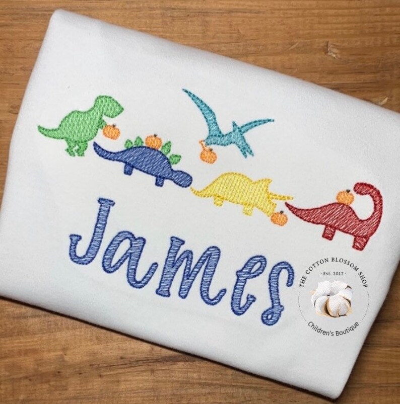May include: A white onesie with a blue embroidered name "James" and five colorful dinosaurs carrying pumpkins. The dinosaurs are green, blue, yellow, and red. The onesie has a small logo in the bottom right corner that says "The Cotton Blossom Shop - Est. 2017 - Children's Boutique."