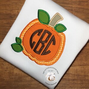 May include: A white t-shirt with an orange pumpkin applique featuring a brown monogram. The pumpkin has white polka dots and green leaves. The t-shirt has a small white label with the text "The Cotton Blossom Shop - Est. 2017 - Children's Boutique".