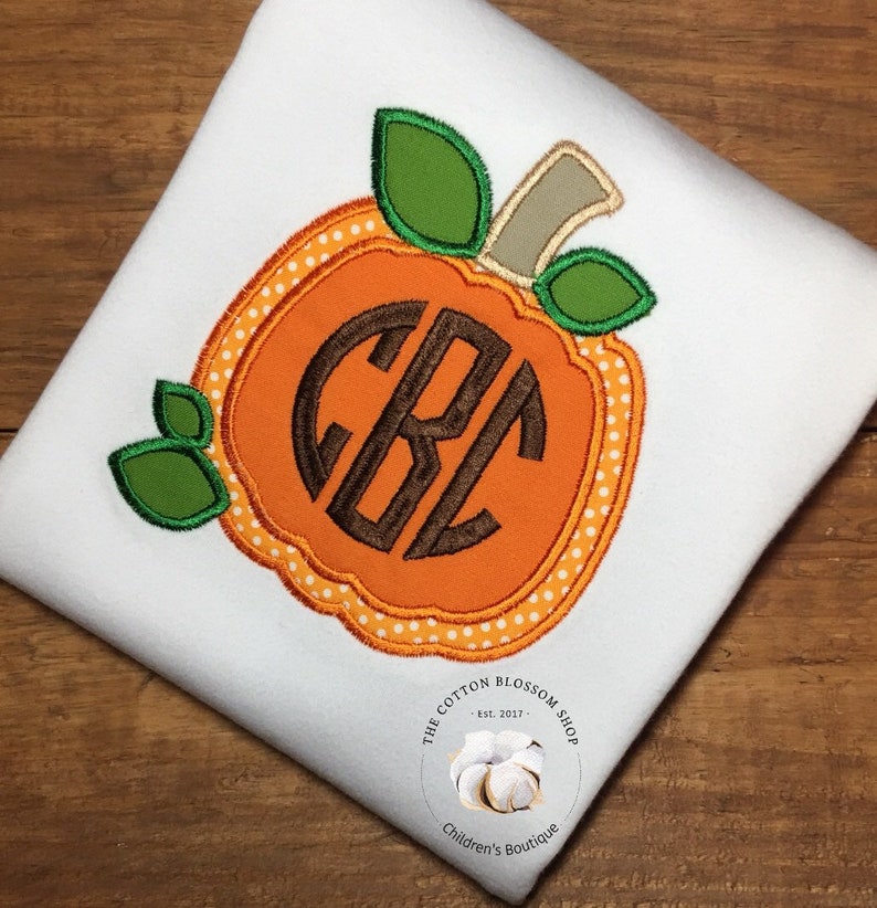 May include: A white t-shirt with an orange pumpkin applique featuring a brown monogram. The pumpkin has white polka dots and green leaves. The t-shirt has a small white label with the text "The Cotton Blossom Shop - Est. 2017 - Children's Boutique".