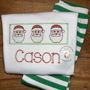 Puede incluir: Un pelele blanco con pantalones a rayas verdes y blancas. El pelele tiene un nombre bordado en rojo "Cason" y una fila de tres caras de Pap&aacute; Noel con gorros rojos y barbas blancas. El pelele tambi&eacute;n tiene un peque&ntilde;o logotipo bordado que dice "The Cotton Blossom Shop Est. 2017 Children's Boutique".