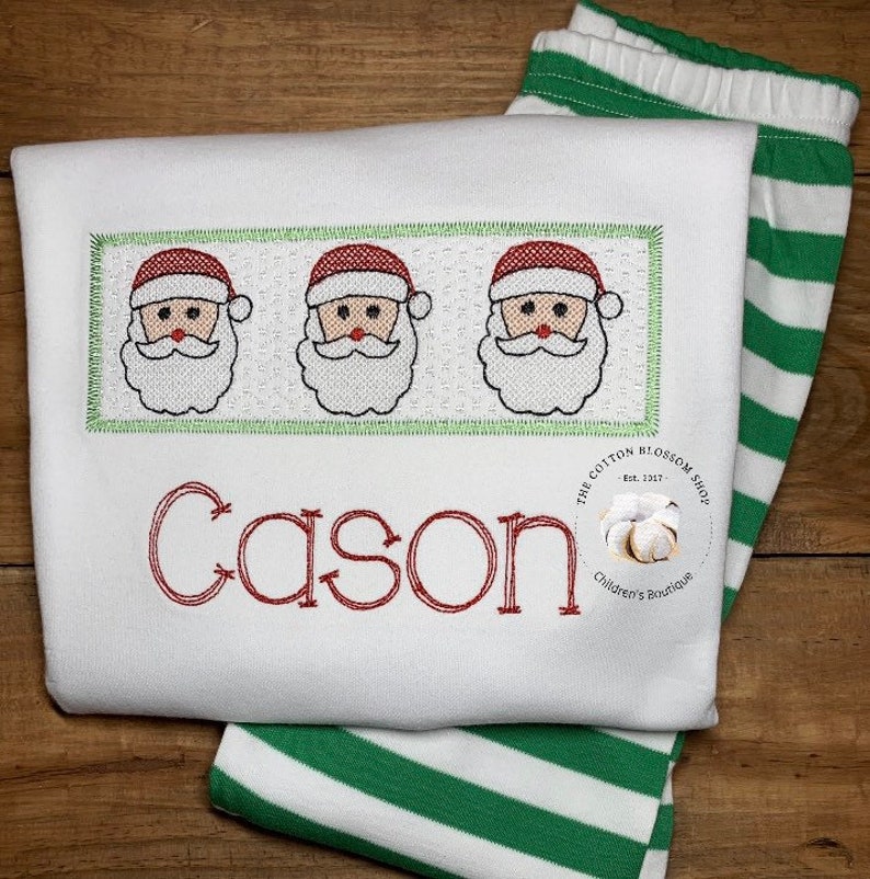 Puede incluir: Un pelele blanco con pantalones a rayas verdes y blancas. El pelele tiene un nombre bordado en rojo "Cason" y una fila de tres caras de Pap&aacute; Noel con gorros rojos y barbas blancas. El pelele tambi&eacute;n tiene un peque&ntilde;o logotipo bordado que dice "The Cotton Blossom Shop Est. 2017 Children's Boutique".