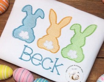 May include: A white onesie with three embroidered blue, orange, and green bunnies. The onesie has the name "Beck" embroidered in blue thread below the bunnies. The onesie is surrounded by colorful Easter eggs.