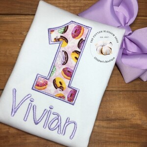 Puede incluir: Un body blanco con un número uno morado bordado en la parte delantera. El número uno está relleno con un patrón de donuts. El nombre Vivian está bordado en morado debajo del número. El body está al lado de un lazo morado.