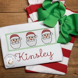 Puede incluir: Tela a rayas blancas y rojas con un lazo verde. Una camisa blanca con un borde verde y tres caras de Pap&aacute; Noel bordadas. La camisa est&aacute; personalizada con el nombre "Kinsley".
