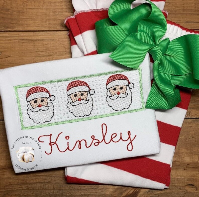 Puede incluir: Tela a rayas blancas y rojas con un lazo verde. Una camisa blanca con un borde verde y tres caras de Pap&aacute; Noel bordadas. La camisa est&aacute; personalizada con el nombre "Kinsley".
