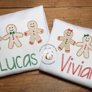 May include: Two white shirts with gingerbread people and names embroidered on them. The shirt on the left has three gingerbread people and the name "Lucas" embroidered in green. The shirt on the right has three gingerbread people and the name "Vivian" embroidered in red. The shirts are on a wooden surface.