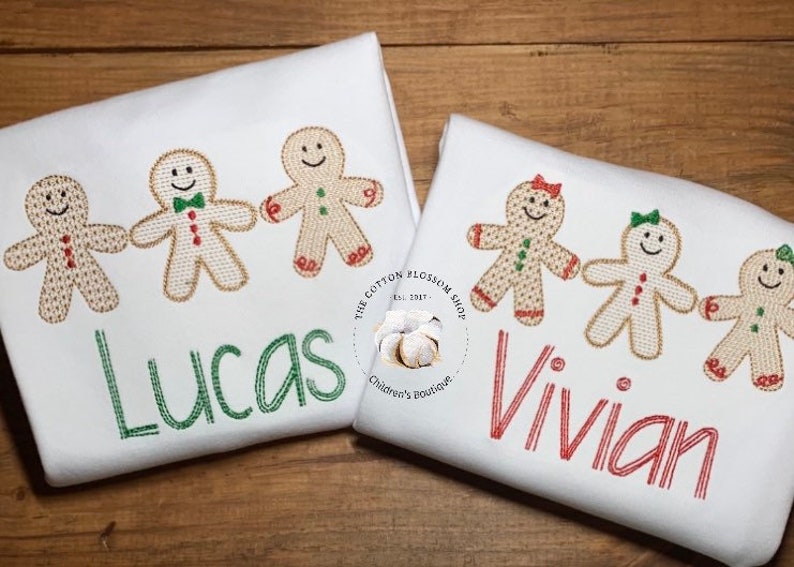May include: Two white shirts with gingerbread people and names embroidered on them. The shirt on the left has three gingerbread people and the name "Lucas" embroidered in green. The shirt on the right has three gingerbread people and the name "Vivian" embroidered in red. The shirts are on a wooden surface.