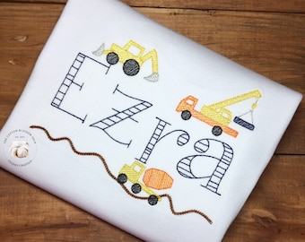 May include: White onesie with the name "Ezra" embroidered in black with yellow construction vehicles around the letters. The vehicles are a dump truck, a crane, and an excavator.