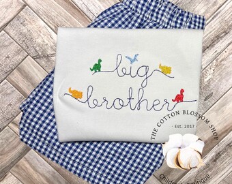 May include: White short-sleeved shirt with blue embroidery that says "big brother" with colourful dinosaur illustrations. The shirt is folded on top of blue and white gingham shorts.