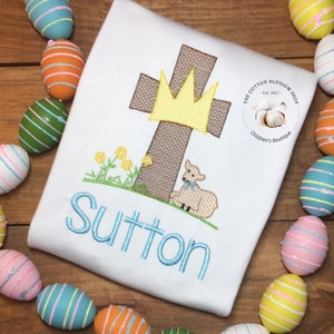 May include: White embroidered baby onesie with a blue embroidered name "Sutton" and a brown cross with a yellow crown and a white lamb. The onesie is surrounded by colourful Easter eggs.