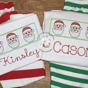 Puede incluir: Dos camisas blancas de manga larga con rayas rojas y verdes. Una camisa tiene una fila de caras de Pap&aacute; Noel bordadas y el nombre "Kinsley" en hilo rojo. La otra camisa tiene una fila de caras de Pap&aacute; Noel bordadas y el nombre "Cason" en hilo rojo.