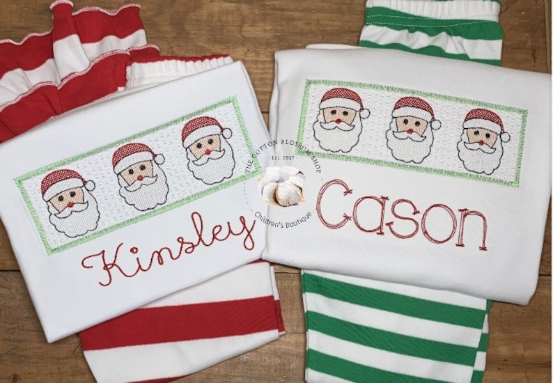 Puede incluir: Dos camisas blancas de manga larga con rayas rojas y verdes. Una camisa tiene una fila de caras de Pap&aacute; Noel bordadas y el nombre "Kinsley" en hilo rojo. La otra camisa tiene una fila de caras de Pap&aacute; Noel bordadas y el nombre "Cason" en hilo rojo.