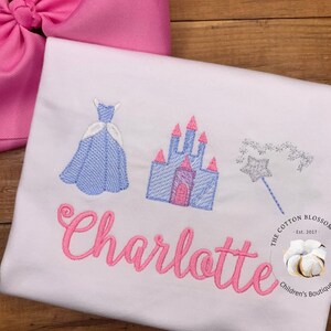 May include: A white t-shirt with pink embroidery of a princess dress, castle, and magic wand. The name "Charlotte" is embroidered in pink below the images. The t-shirt has a small logo in the bottom right corner that reads "The Cotton Blossom Shop Est. 2017 Children's Boutique."