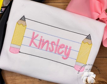 Könnte beinhalten: Weißes personalisiertes T-Shirt mit einem pinkfarbenen und gelben Bleistiftdesign und dem Namen "Kinsley" auf einem blau linierten Papierhintergrund.