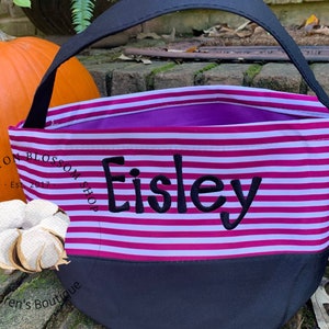 May include: Pink and white striped Halloween trick-or-treat bag with black handles and a black bottom. The bag is personalized with the name "Eisley" in black embroidery. The bag is sitting on a wooden surface next to a pumpkin.