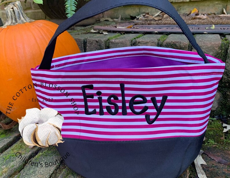 May include: Pink and white striped Halloween trick-or-treat bag with black handles and a black bottom. The bag is personalized with the name "Eisley" in black embroidery. The bag is sitting on a wooden surface next to a pumpkin.